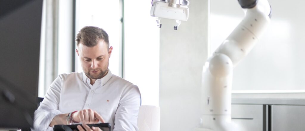 Mann im modernen Büro mit Tablet und Roboterarm. Ein lächelnder Mitarbeiter in weißem Hemd sitzt auf einem Bürostuhl und hält ein Tablet, während ein großer, weißer Roboterarm neben ihm zu sehen ist. Ideal für Inhalte über moderne Technologie, Automatisierung und Arbeitsplatzinnovationen.