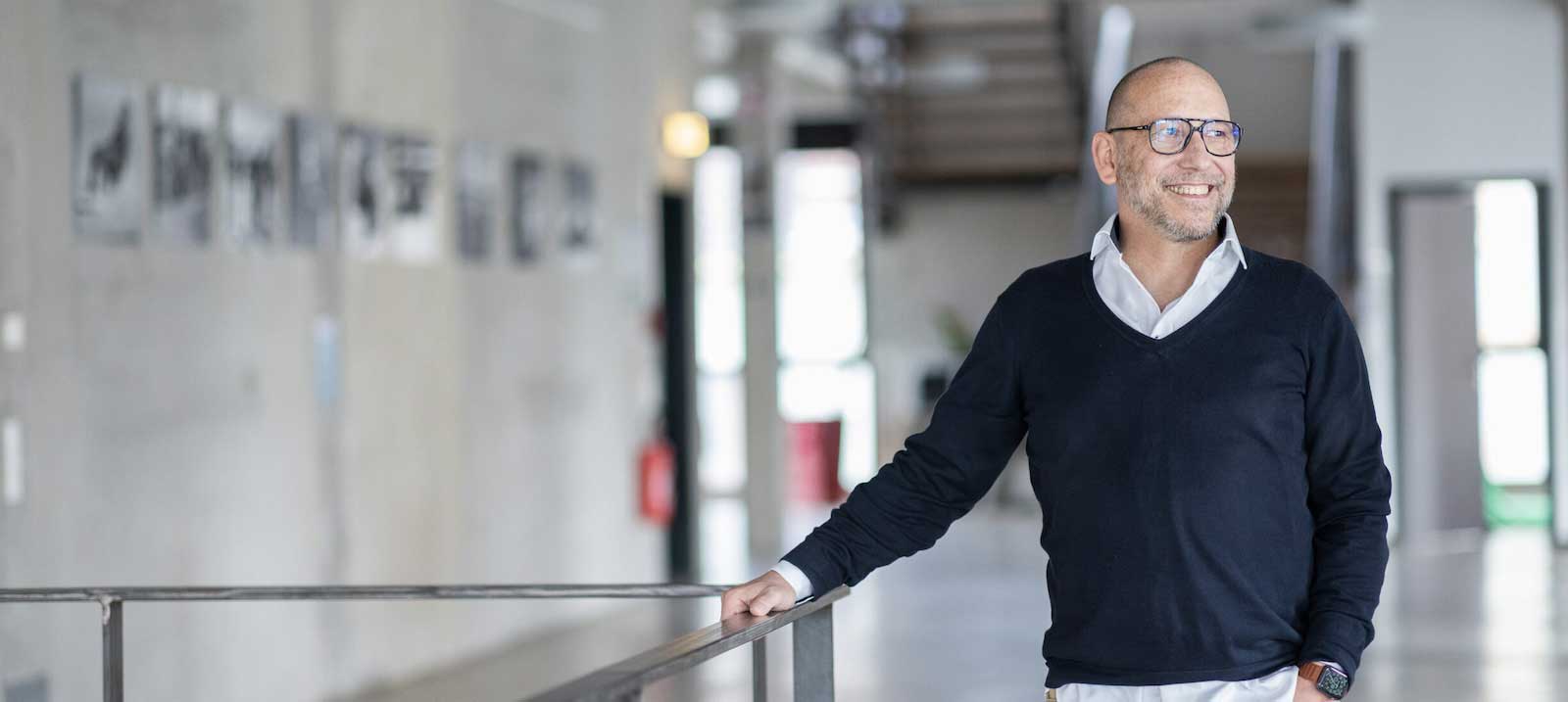 Geschäftsführer Andreas Bader von Objective Partner lehnt lächelnd an einem Geländer in einem modernen Bürogebäude, gekleidet in einem dunklen Pullover über einem weißen Hemd, mit einer Brille.