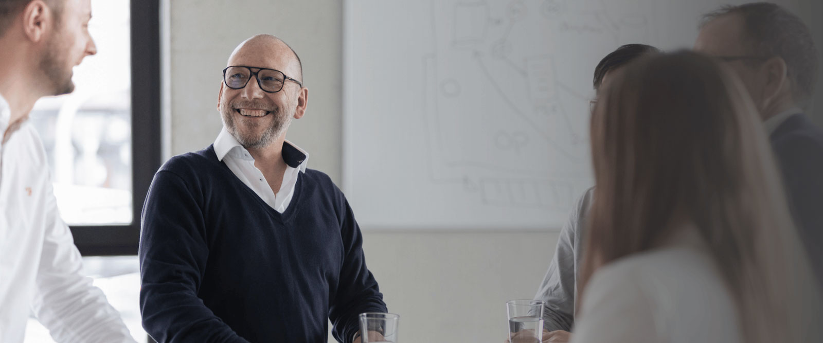 Geschäftsführer Andreas Bader von Objective Partner lächelt während einer Besprechung im Büro, umgeben von Teammitgliedern in einer hellen, modern gestalteten Umgebung.